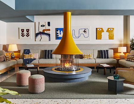 A bright, modern lounge with a circular fireplace, mustard-yellow centerpiece cone, surrounding beige sofas, colorful abstract art, and cozy ottomans, all under soft lighting.
