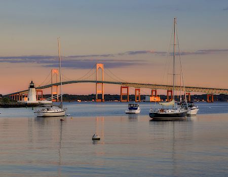 Sailboats drift on calm water with a bridge and lighthouse in the distance, as a warm sunset washes the scene in soft golden light.