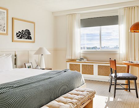 A bright hotel room with a bed, striped duvet, bench at the foot, desk and chair by a window, warm lighting, and simple wall art.