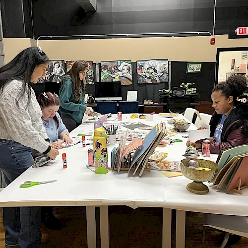 A group of kids and adults create art at a long table in a bright studio, with notebooks, paints, and colorful crafts spread out, collaborating together.