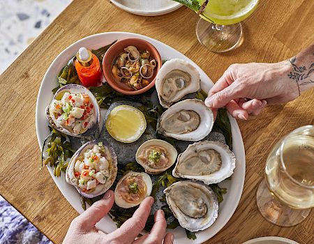 A table with a seafood spread: oysters on the half shell, small bites in colorful cups, a lemon, and drinks on a wooden table, diners&rsquo; hands reaching in.