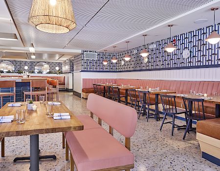 A modern, airy eatery with pink booths, wooden tables, blue-and-white patterned walls, pendant lights, and a mix of booth and table seating.