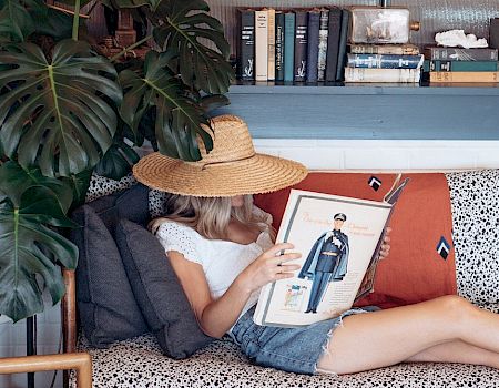 A person in a sunhat sits on a sofa reading a magazine or book, surrounded by a plant, books, and a blue shelf, cozy and relaxed.