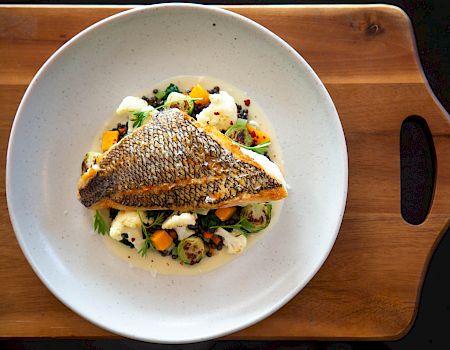 Crispy grilled fish atop a bed of colorful vegetables and creamy sauce, served on a white plate placed on a wooden board.