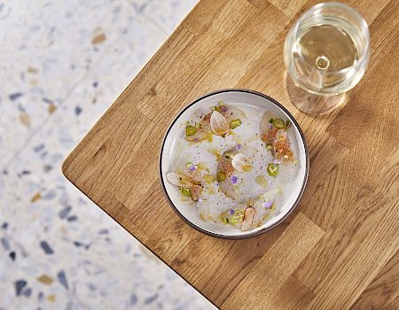 A bowl of cloudy soup with white broth, small fried dumplings or croutons, and a few pinkish toppings, next to a glass on a wooden table.