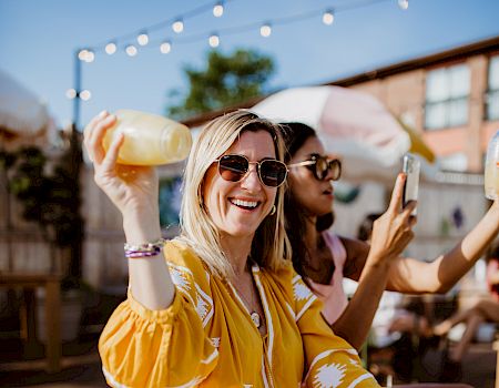 Two friends at an outdoor hangout, one in a yellow dress raising a drink and smiling at the camera, string lights above.