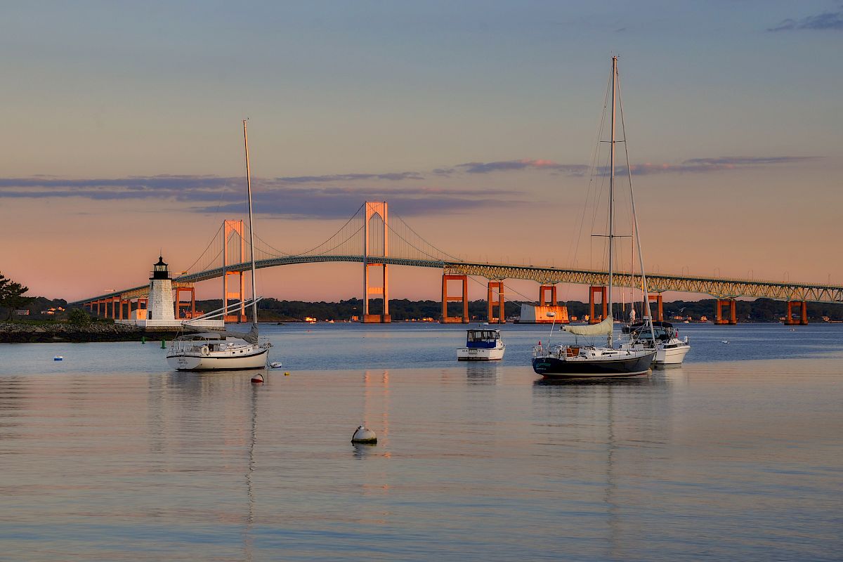 Sailboats rest on calm water with a long bridge and lighthouse at sunset, reflecting warm colors across the tranquil harbor.