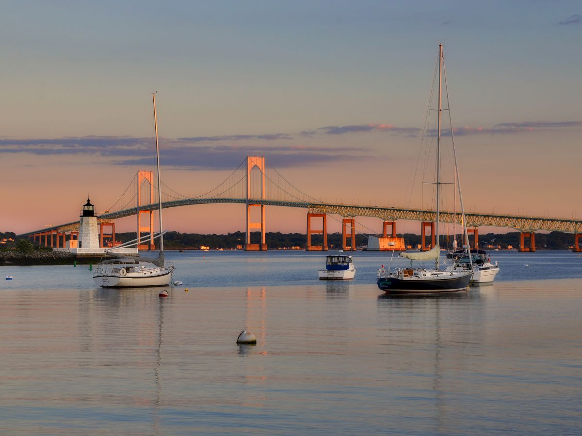 Sailboats rest on calm water with a long bridge and lighthouse at sunset, reflecting warm colors across the tranquil harbor.