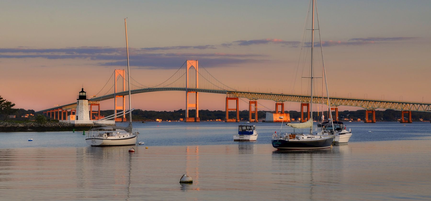 Sailboats rest on calm water with a long bridge and lighthouse at sunset, reflecting warm colors across the tranquil harbor.
