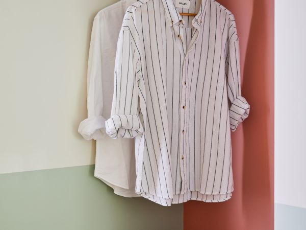 Two striped button-up shirts hang in a closet, with a hat and stacked books on a shelf below, set against a pastel color-blocked wall.