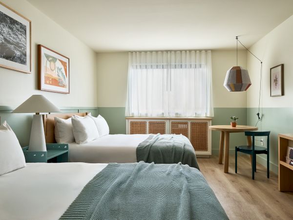 A bright hotel room with two neatly made twin beds, teal bed runners, wall art, a window with sheer curtains, and a desk area with a chair, TV, and warm wood accents.