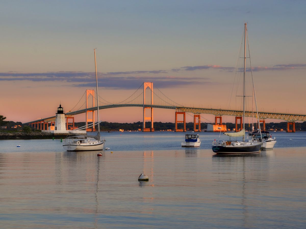 A calm coastal scene at sunset with sailboats anchored in still water and a distant suspension bridge lit by warm orange-pink light.