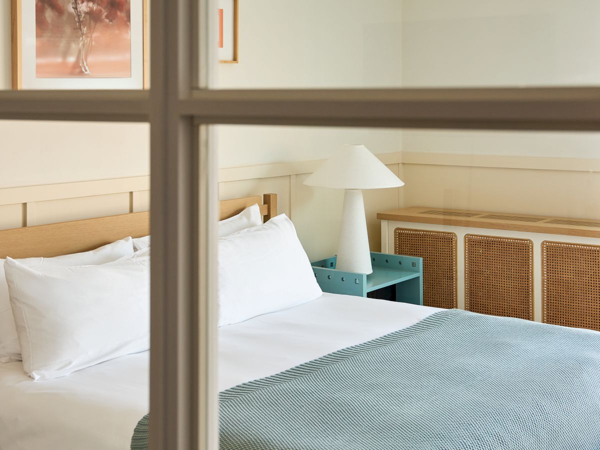 A cozy hotel bedroom with a neatly made bed, white pillows, a teal bed runner, a bedside table and lamp, viewed through a windowed wall.