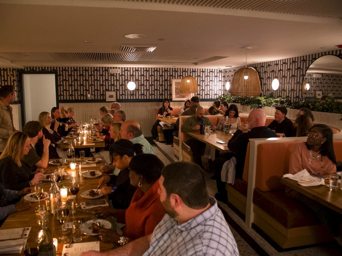 A crowded restaurant with people dining at long booths and tables, warm lighting, and a mosaic-tiled wall in the background.