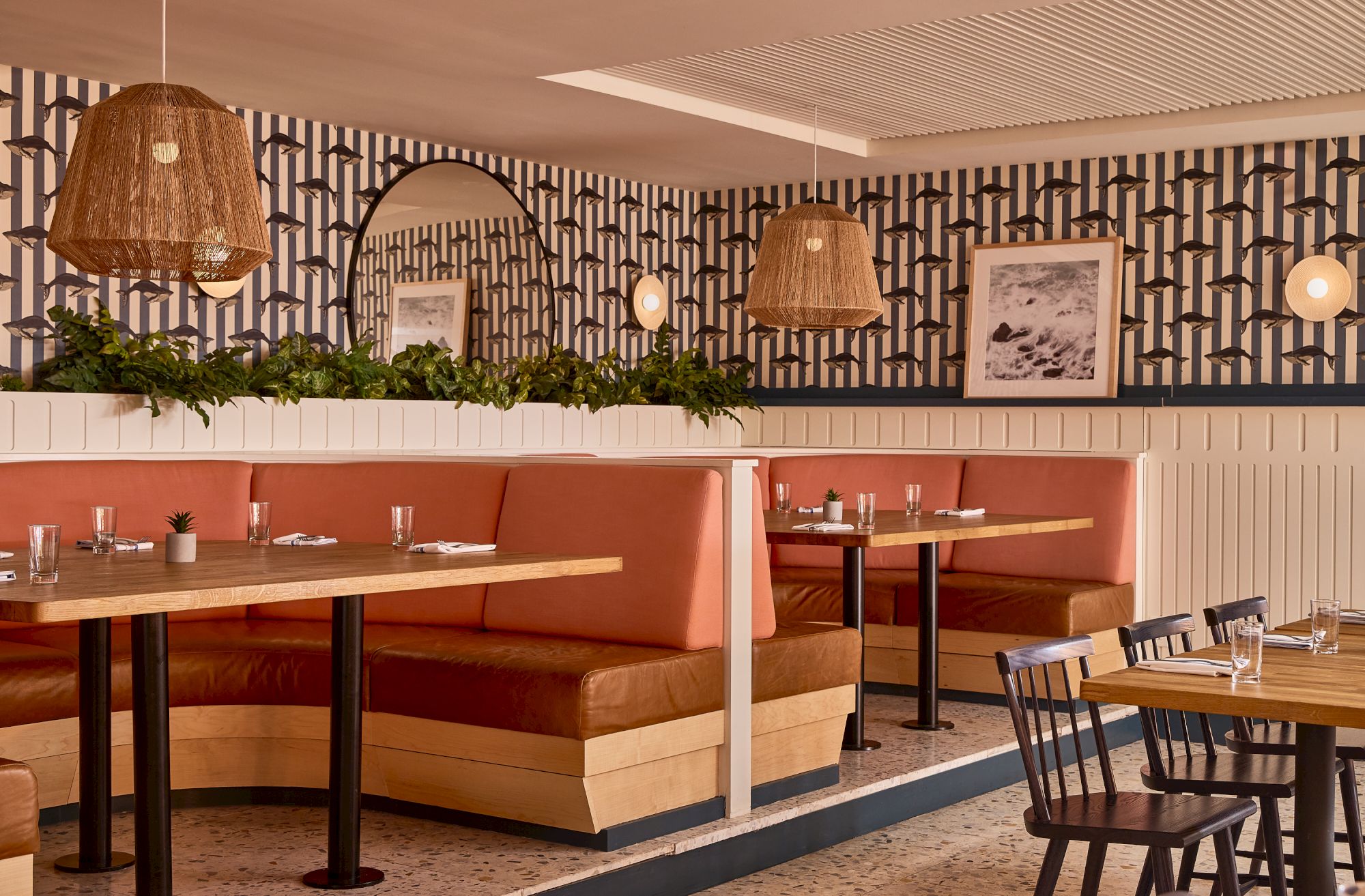 A stylish dining area with pink booth seating, wooden tables, black chairs, pendant lights, patterned wallpaper, and framed art.