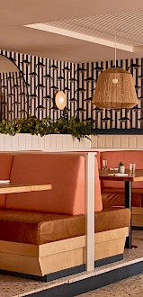A stylish restaurant interior with peach-banquette seating, wooden tables, woven pendant lights, black-and-white geometric wallpaper, and green plants.