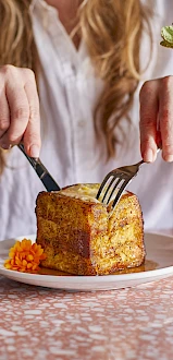 A person in a white shirt slices a savory-looking square dinner roll or cake on a plate with carrot side, beside a bright orange cocktail.