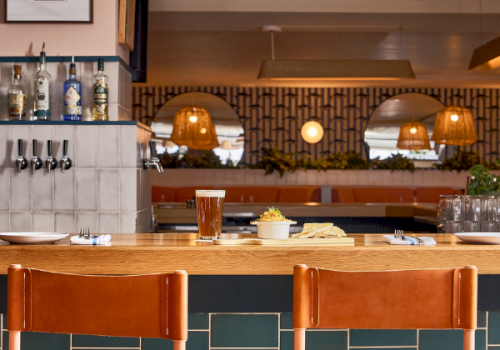 A bar counter with two wooden stools, glassware, and a row of bottles behind a tiled backsplash, warm lighting, and a cozy restaurant vibe.