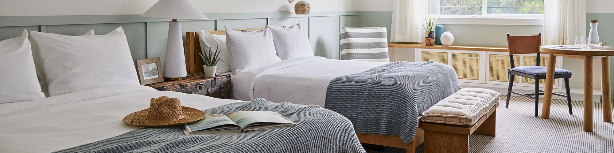 The image shows a cozy hotel room with two beds, a wooden table with chairs, a bench, a hat, an open book, plants, and framed pictures on the wall.