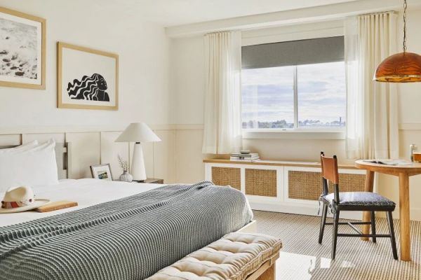 A bright, modern bedroom with a bed, desk, chair, and window. Decor includes framed artwork, a table lamp, and a woven bench at the foot of the bed.