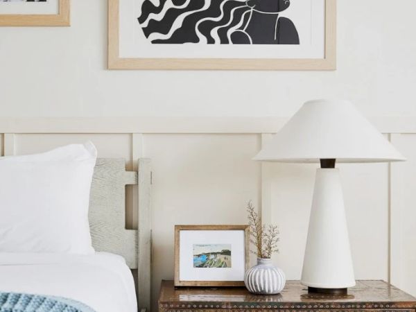 A minimalist bedroom scene with a bed, nightstand, table lamp, small framed picture, vase, and art on the wall above.