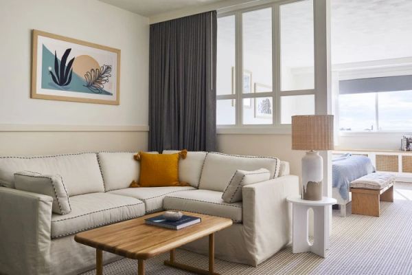 A cozy living space with a white sectional sofa, wooden coffee table, abstract wall art, lamp, and large window with gray curtains. Bed visible in back.