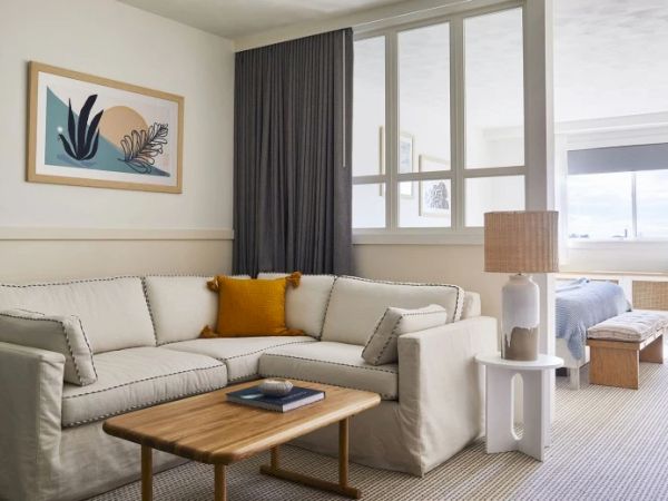 A cozy living space with a white sectional sofa, wooden coffee table, abstract wall art, lamp, and large window with gray curtains. Bed visible in back.