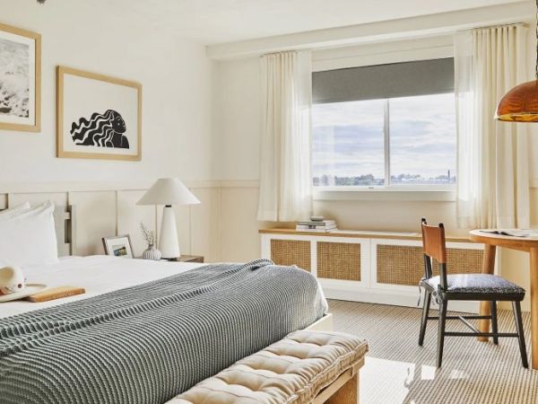 The image shows a bright, cozy bedroom with a large bed, a wooden desk and chair, artworks on the wall, and a window with a scenic view outside.