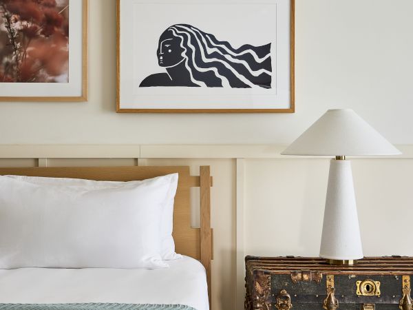 A cozy bedroom with a framed artwork of a black-and-white profile and wavy hair above a vintage trunk beside a white lamp, next to a neatly made bed.