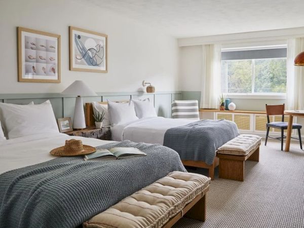 This image shows a cozy hotel room with two neatly made beds, a window, artwork on the wall, a small table with chairs, and modern decor elements.