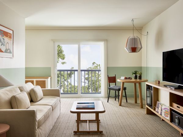 A cozy living room with a beige sofa, wooden coffee table, TV stand, a small dining area by a sliding door balcony, and soft natural light.