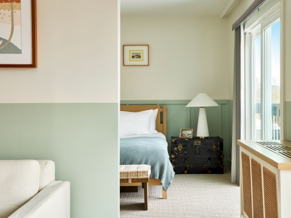A tidy hotel room with a bed, nightstand, lamp, and dresser visible through a doorway from a living area, framed wall art, and soft natural light.