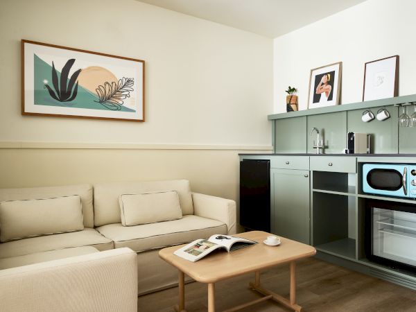 A compact, modern living area with a beige sofa, a wooden coffee table, and a compact kitchenette featuring mint cabinets, a microwave, and an oven, with framed art on the wall.