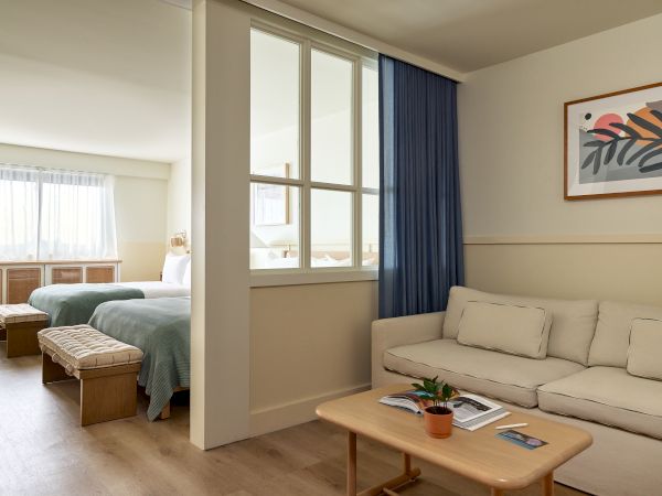 A bright hotel suite with a living area and a separate bedroom, featuring a beige sofa, wooden coffee table, blue curtains, and beds in the background, all in a clean, airy space.