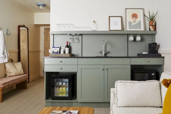 The image shows a home kitchenette with a sink, shelves with glassware, and appliances. There’s a couch and a coffee table in the foreground.