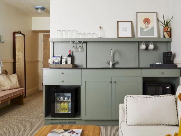 The image shows a home kitchenette with a sink, shelves with glassware, and appliances. There’s a couch and a coffee table in the foreground.