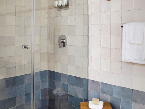 This image shows a tiled shower area with glass doors, a wall-mounted towel rack with a towel, and a wooden stool with soap on it.