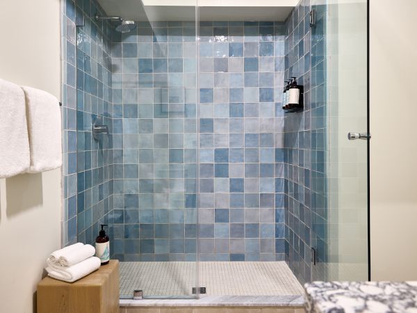 A glass-front shower with blue square tiles, a small bench area, stacked white towels, and a wooden counter with soap dispenser inside the stall.