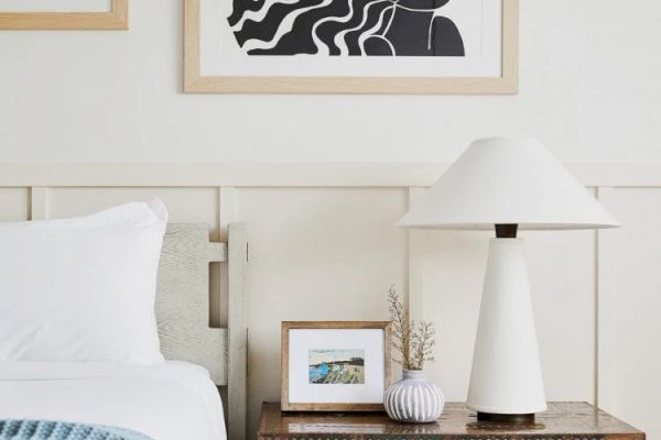 A cozy bedroom with a bedside table holding a white lamp, a framed photo, and a small vase. Above hangs an abstract black and white artwork.