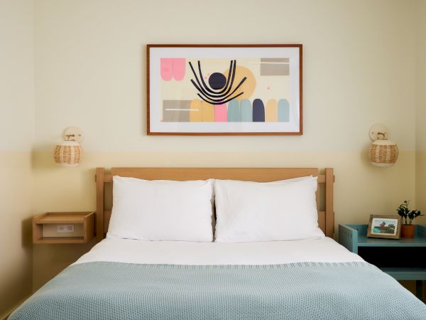 A neatly made bed with white pillows and a light blue duvet, framed abstract art above, and matching bedside tables with lamps on each side.