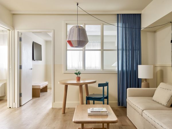 A bright, modern living area with a beige sofa, wooden coffee table, round dining table, blue curtains, wall lamp, and a view into a small bedroom; clean, airy decor.