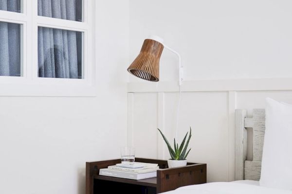 This image shows a minimalist bedroom with a wooden nightstand, a plant, books, a glass of water, and a wall-mounted bamboo lamp.