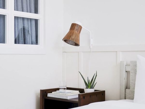 This image shows a minimalist bedroom with a wooden nightstand, a plant, books, a glass of water, and a wall-mounted bamboo lamp.