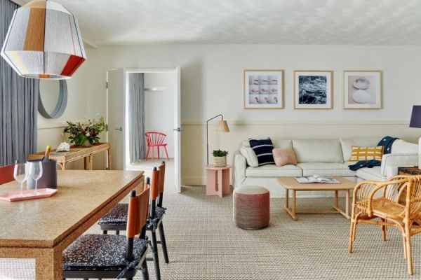 A modern living room with a sofa, rattan chair, framed wall art, and a dining area featuring a wooden table, pendant light, and upholstered chairs.