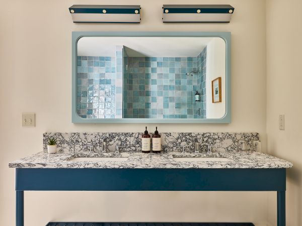 A bathroom vanity with a speckled granite counter, blue trim, a rectangular mirror, and turquoise mosaic tiled shower reflected&mdash;simple and serene.