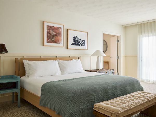 A cozy hotel room with a king bed, teal bedside table, two wall art prints, a tufted bench at the foot, and a large window with sheer curtains.