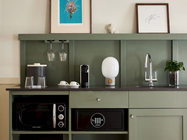 A compact kitchenette with a muted green cabinet, mini appliances, a sink, and framed art above; a cozy, studio-style setup.