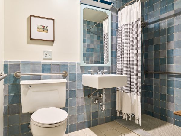 A small blue-tiled bathroom with a wall-mounted sink, mirror, toilet, towel rail, and a striped shower curtain, all in a compact space.