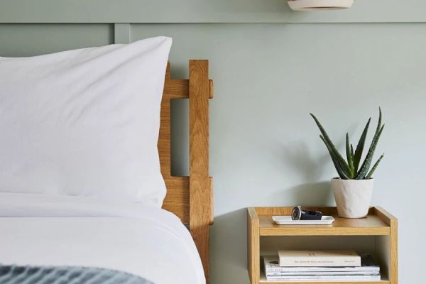 A bedside table with books, a potted plant, and a phone. Next to it is a bed with white bedding and a wooden headboard.