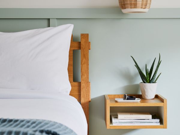 A cozy bedroom with a light wooden bed frame, white bedding, a blue-gray blanket, a small wall shelf with books, a potted plant, and a wicker wall light above.